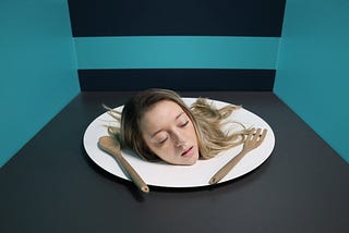 image: woman’s head on a white dinner plate between a spoon (left) and fork (right); the background is black and teal.