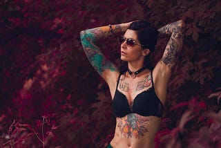 Standing woman wearing sunglasses with bright tattoos on her arms torso and upper chest.