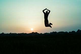 Black silhouette of a young man jumping up off the ground celebrating happiness with an orange, pink, yellowish setting sun in a blue sky. The image was chose by Spiritual Lifestyle Writer Christine McDonald to show the joy one feels when they allow the Spirit within them to guide them through life’s struggles successfully.