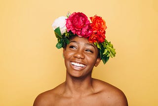 A shoulders-up view of a person with a flower crown, smiling, in front of a yellow background.