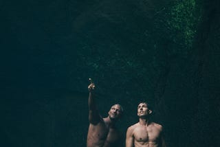 Two men in swim suits wade through shallow water.