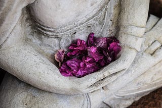 Stone statue shown seated in lotus pose from the chest down, hold magental petals in its lap