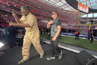 A man with dark skin signing a song next to a man with light skin wearing glasses. They are performing at the end zone at the Super Bowl.