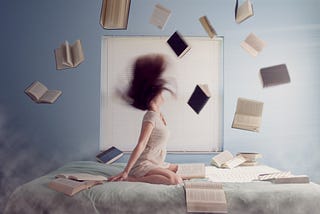 Woman kneeling on bed with multiple books in the air around her. Her long hair is captured being thrown back around her head.