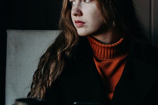 A woman with long brown hair and wearing a jumper and jacket, gazes out a window. She is holding a black phone and a coffee cup sits on the table in front of her.