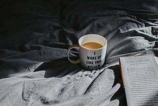 photo of a coffee mug that reads “I woke up like this #tired.” The mug is sitting on gray sheets beside an open book.