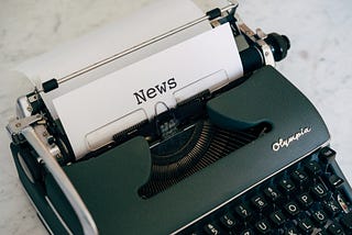 Dark green modern typewriter with a piece of paper in it that has the word “news” typed on it in typewriter font