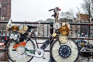 Bicycle covered with clocks, parked on a low bridge over a canal