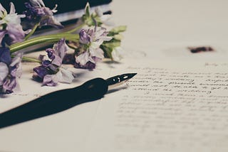 A black fountain pen sitting on a letter; next to it, a sprig of purple flowers rests.