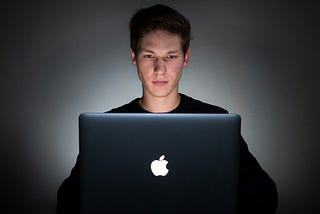 A young man is lit by the glow of his laptop in the dark.