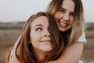 Two young women with one smiling and hugging from behind. The second woman is looking up in adoration.