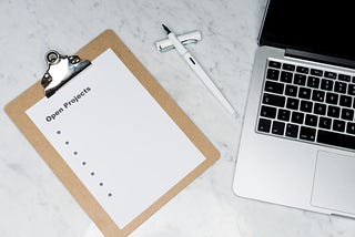 Clipboard with a piece of paper reading ‘open projects’ ready for a list, with a pen and a laptop on a desk