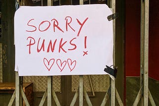 Handwritten poster taped to metal bars of a gate. Poster reads, “Sorry Punks” with three lovehearts drawn underneath.