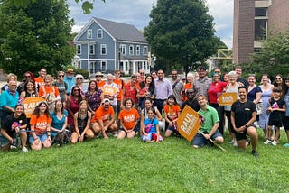 Around 50 people standing outside by the Brookline Library, near School Street. They are posing together for a group photo; many of them are wearing orange Raul t-shirts and/or holding orange Raul campaign signs.