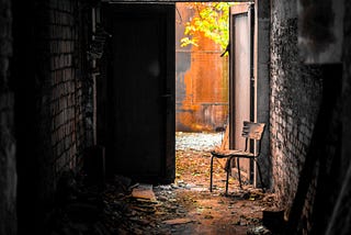 A chair sits in the open door at the end of a dark enclosed alley that promises light outside.