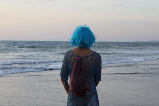 A woman with blue hair staring out into the ocean admiring its calm scenic beauty.