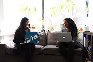 Two women of color smile at each other with open laptops.