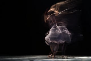 A teenage girl dances barefoot on a dark stage, her form photographed blurry from the movement.