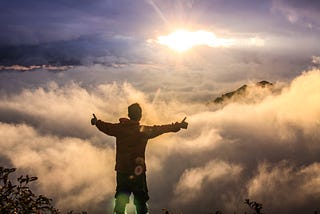 Man standing over a cloud filled valley, the sun shining over top and he’s giving it two thumbs up.