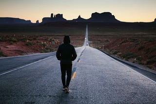 A hooded figure walks along the yellow, dashed line in the center of a road going up into the mountains on the horizon.
