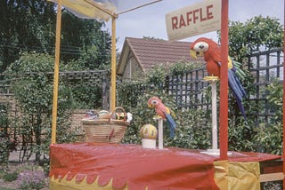 A stall with a sign saying ‘raffle’ on it. The stall is covered in red and yellow material and is situated in a garden or other green space. There is a basket with what looks like small prizes in sitting on the stall as well as two brightly coloured statues of parrots on perches and a yellow ball.