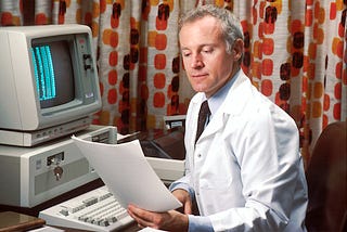A middle-aged man wearing a white medical coat sits in front of a computer reading notes that he’s holding in his left hand.