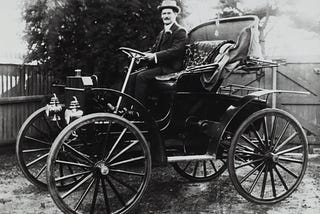 An original horseless carriage in black and white with a man in a hat sitting in the driver’s seat