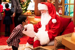A young boy in a plaid shirt leaning forward to a sitting down Santa Claus who’s holding a letter in his left hand.