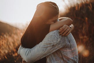 Two young people embrace in a field of grass beneath the glow of late afternoon sun.