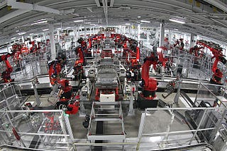A large, Tesla factory floor is depicted, showcasing an automated assembly line. Numerous red robotic arms are stationed along the assembly line, engaged in various tasks. The area is enclosed by clear safety barriers and the floor is clean and well-lit by overhead lighting. In the center of the image, a partially assembled vehicle is visible, indicating that the factory specializes in automobile manufacturing.