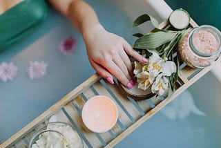 Uma cena serena de um spa mostra uma mão descansando em um suporte de madeira sobre uma banheira. No suporte, encontram-se elementos típicos de uma experiência de spa, como um pote de sais de banho cor-de-rosa, uma vela aromática suave, rodelas de pepino fresco, e flores brancas delicadas. A água azulada da banheira está adornada com pétalas de flores, sugerindo um ambiente relaxante e luxuoso, ideal para um momento de autocuidado profundo.