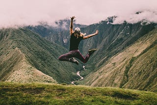 Person with long hair in athletic wear photographed jumping up in a celebratory fashion on top of a mountain.