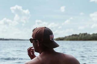 Shirtless man in a hat looks across a lake