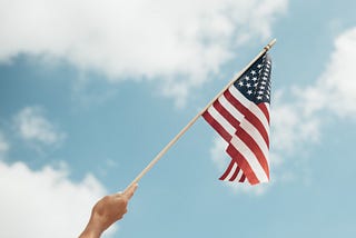 American flag being held up against a blue, cloud-filled sky.