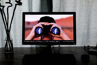 The image shows a computer screen displaying a man looking through binoculars. Instead of traditional lenses, the binoculars have Facebook logos on each side, symbolizing the concept of viewing or spying through Facebook. The setup is on a desk with a keyboard and a vase of decorative branches placed to the left of the monitor. The background behind the monitor consists of light-colored curtains, creating a simple and minimalistic setting.