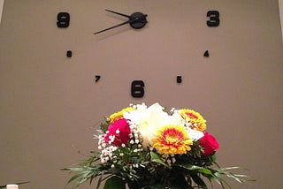 A unique wall clock. In front of it, is a table and chairs with a bouquet of flowers.