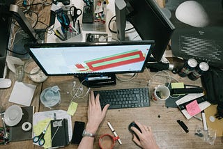 A very messy workspace is shown from above, with a keyboard, monitor, smartphone, earbuds, cords, sticky notes, scissors, several half-empty mugs, and general disarray.