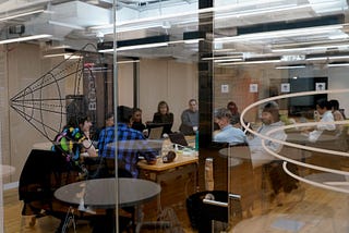 A behind-the-scenes image of our board of directors and advisors, staff, and community members at NYU ITP. The image is taken in front of a glass door. Inside the room, around 10 people gathered by a table, working on their laptops.