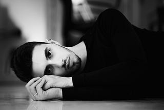 Black and white image of a man lying down on the floor, hands clasped together.