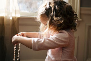 A side-view of a little girl, barely the height of the windowsill, wearing a pink dress holding out a string of pearls as if determining to put them around her neck. Her hair short and wavy but maturely coiffed like a woman’s but with a barrette. A representative photo of a girl growing up too fast against more idealistic fashion.