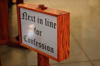 The words Next in line for Confession appear on a sign that is framed in faux wood. The sign is held up by a stake and it appears in front of a walkway