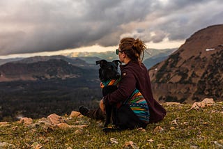 A woman hugging her dog and watching a storm roll in while her dog checks for activity behind them.