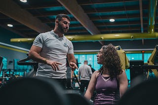 A lady in a gym, wearing a purple exercise shirt, listening to her personal trainer, who is standing up, looking and speaking to her.