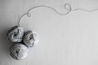 Three balls of light grey yarn with a singular thread looping at the top on a grey background