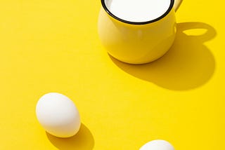 Two white eggs sit in front of a cup of milk.