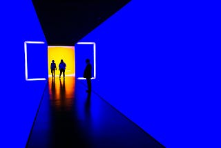 Three people standing in a futuristic museum-like place with a yellow background and in between two blue walls