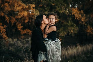 Portrait of mother kissing her young child in front of autumn trees