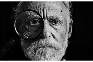 Old man with a questioning look on his face holding a magnifying glass to his right eye.