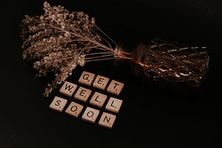 Vase of flowers on its side with scrabble letters below that say “Get well soon”.