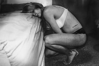 A woman in her undergarments kneeling beside a bed, rests her head on the mattress in defeat.